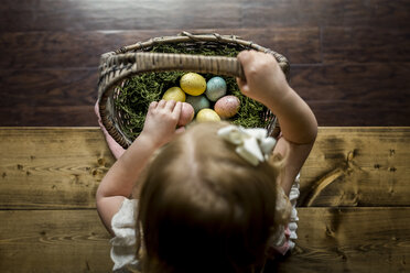 Overhead view of girl with basket of Easter eggs sitting on wooden table at home - CAVF32364