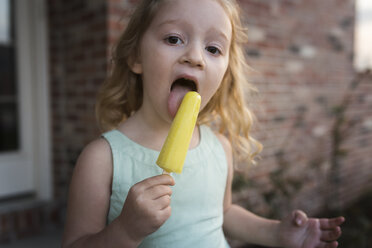 Portrait of girl eating popsicle stick while standing against house - CAVF32367