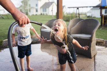 Cropped hand of man holding hose while girl drinking water and brother standing beside her at yard - CAVF32368