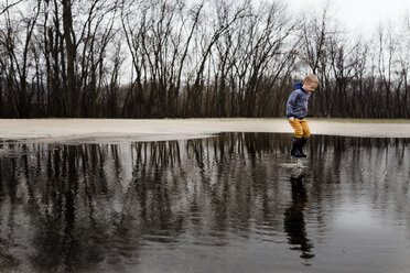 Full length boy jumping in puddle against bare trees - CAVF32380