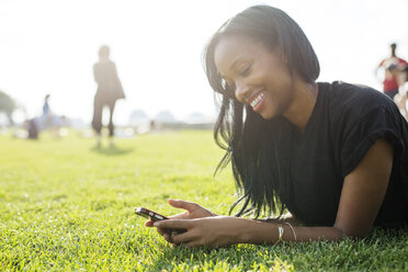 Glückliche Frau, die auf einem Feld liegend eine SMS schreibt - CAVF32430