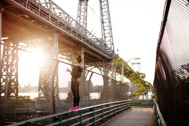 Woman standing on railing while looking at Williamsburg Bridge on sunny day - CAVF32448