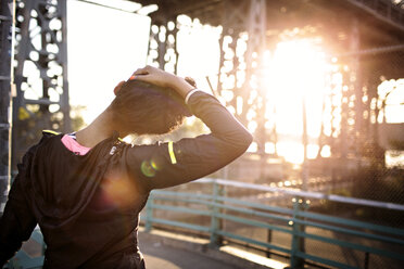 Rückansicht einer Joggerin, die ihren Nacken auf einer Fußgängerbrücke streckt, an einem sonnigen Tag - CAVF32450