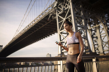 Sporty woman listening to music while standing against Williamsburg Bridge - CAVF32453