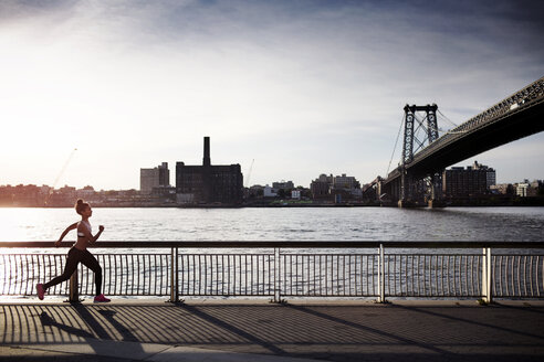 Entschlossene Frau joggt auf der Promenade mit der Williamsburg Bridge im Hintergrund - CAVF32454