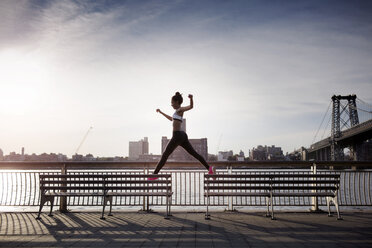 Sportliche Frau springt auf Bänken mit Williamsburg Bridge im Hintergrund - CAVF32455