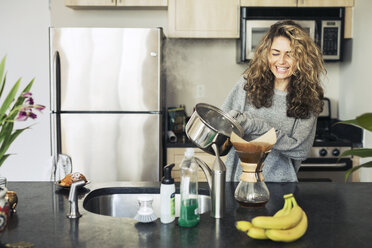 Glückliche Frau gießt Kaffee in einen Filter am Küchentisch - CAVF32476