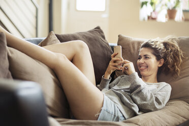 Glückliche Frau schreibt eine SMS, während sie zu Hause auf dem Sofa liegt - CAVF32483