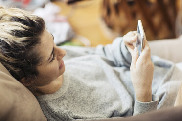 Draufsicht auf eine Frau, die ein Telefon benutzt, während sie zu Hause auf dem Sofa liegt - CAVF32485