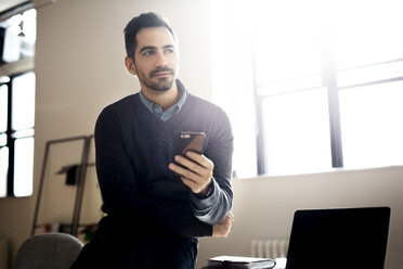 Thoughtful businessman holding smart phone and sitting in office - CAVF32526