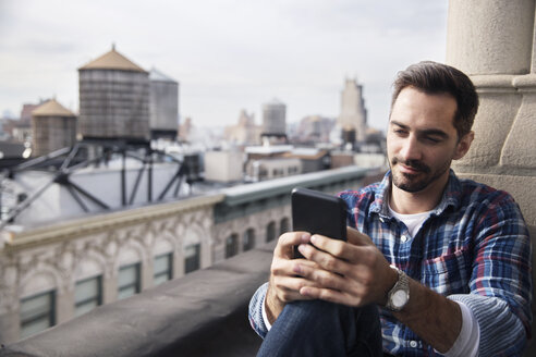 Lächelnder Mann, der sein Smartphone benutzt, während er auf dem Balkon gegen den Himmel sitzt - CAVF32553