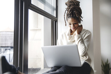 Smiling businesswoman using laptop while sitting at window sill in office - CAVF32555