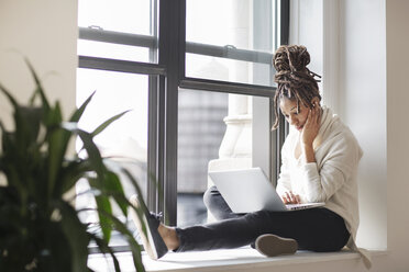 Geschäftsfrau arbeitet mit Laptop, während sie am Fenster im Büro sitzt - CAVF32556