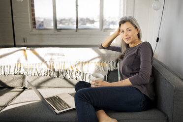 Portrait of smiling woman holding coffee mug and sitting by laptop on sofa - CAVF32599