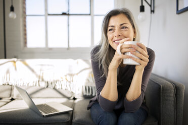 Smiling woman holding coffee mug while sitting by laptop on sofa in living room - CAVF32600