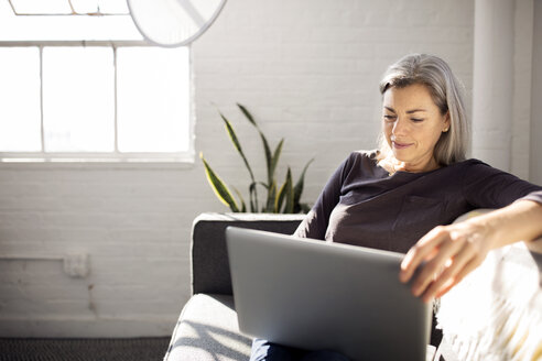 Frau, die einen Laptop benutzt, während sie zu Hause auf dem Sofa im Wohnzimmer sitzt - CAVF32601