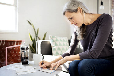 Concentrated woman writing on paper at table in living room - CAVF32604