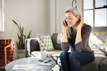 Woman talking on phone while sitting on sofa in living room at home - CAVF32605