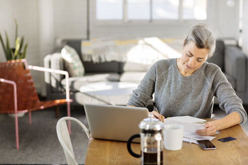 Konzentrierte Frau schreibt auf Papier, während sie im Wohnzimmer am Laptop arbeitet - CAVF32615