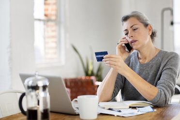 Woman holding debit card while using phone at home - CAVF32617