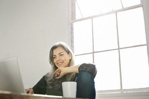 Lächelnde Frau mit Laptop am Tisch vor dem Fenster zu Hause - CAVF32625