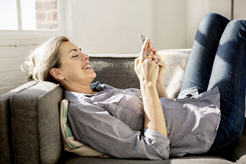 Happy woman using smart phone while lying on sofa at home - CAVF32633