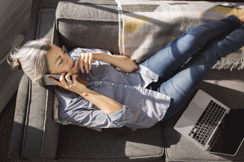 Woman talking on smart phone while lying by laptop on sofa at home - CAVF32635