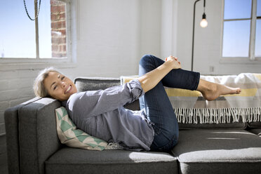 Porträt einer glücklichen Frau, die ihre Knie umarmt, während sie zu Hause auf dem Sofa liegt - CAVF32637