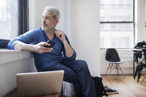 Thoughtful mature man holding smart phone while looking through window at home - CAVF32665