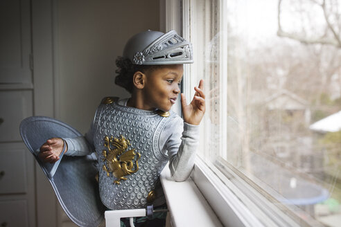 Curious boy dressed up in armor costume looking out through window at home - CAVF32679