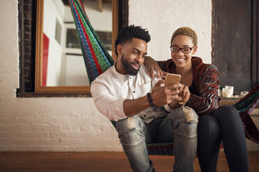 Happy young couple using smart phone while sitting on hammock at home - CAVF32730