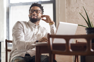 Happy man looking away while using laptop at table - CAVF32742