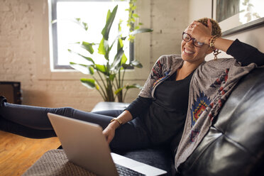 Cheerful woman using laptop on sofa at home - CAVF32750