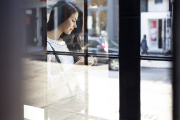 Junge Frau mit Smartphone durch Glaswand gesehen - CAVF32782