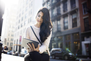 Schöne Frau, die einen Tablet-Computer benutzt, während sie an einem sonnigen Tag auf einer Straße in der Stadt sitzt - CAVF32790