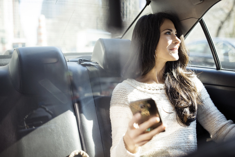 Nachdenkliche Frau, die ihr Smartphone in der Hand hält und durch das Taxifenster schaut, lizenzfreies Stockfoto