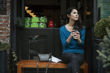 Nachdenkliche Frau hält ein Einwegglas, während sie auf einer Bank vor einem Café sitzt - CAVF32818
