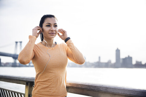 Sportliche Frau mit Kopfhörern am Fluss mit der Williamsburg Bridge im Hintergrund - CAVF32846
