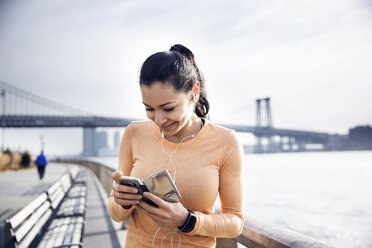 Happy female athlete listening music through smart phone with Williamsburg Bridge in background - CAVF32847