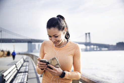 Glückliche Sportlerin hört Musik über ihr Smartphone mit der Williamsburg Bridge im Hintergrund - CAVF32847