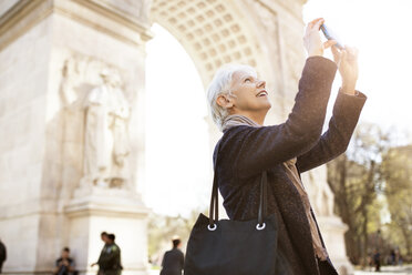 Side view of senior woman photographing through smart phone while standing against triumphal arch in city - CAVF32889