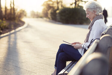 Ältere Frau benutzt einen Tablet-Computer, während sie auf einer Bank am Fußweg sitzt - CAVF32902