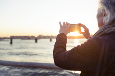 Rückansicht einer älteren Frau, die bei Sonnenuntergang am Meer fotografiert - CAVF32908