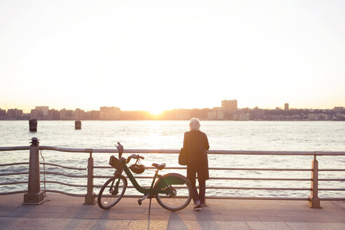 Rückansicht einer älteren Frau, die während des Sonnenuntergangs mit dem Fahrrad am Meer steht - CAVF32913