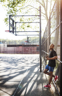 Mann hält Handy in der Hand und lehnt sich an den Zaun eines Basketballplatzes - CAVF32946