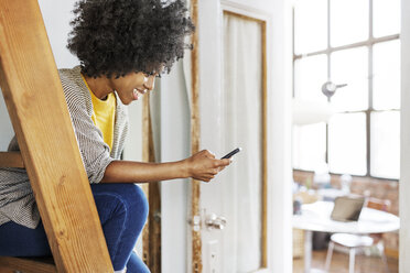 Glückliche Frau, die ein Mobiltelefon benutzt, während sie zu Hause auf einer Leiter sitzt - CAVF32995