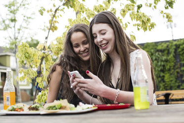 Smiling female friends using smart phone while sitting at sidewalk cafe - CAVF33020