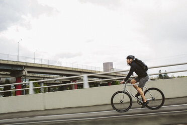 Seitenansicht eines männlichen Pendlers mit Rucksack auf dem Fahrrad auf einer Brücke gegen den Himmel - CAVF33065