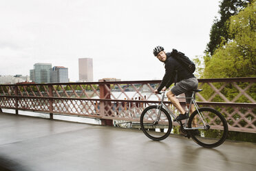 Seitenansicht eines männlichen Pendlers, der auf einer Brücke über einen Fluss gegen den Himmel Fahrrad fährt - CAVF33066