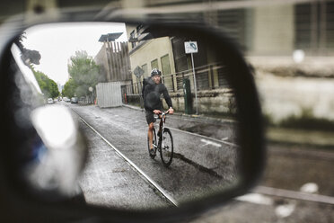 Männlicher Pendler, der auf einer nassen Straße Fahrrad fährt, gesehen durch den Seitenspiegel eines Autos - CAVF33072